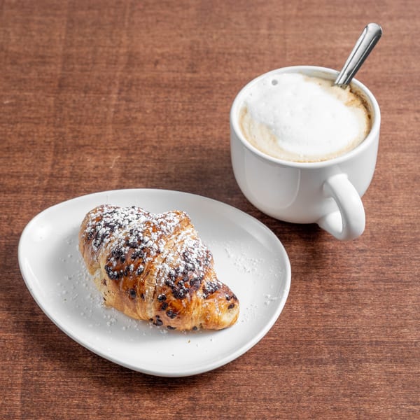 Chocolate chip croissant with powdered sugar on a white plate next to a foamy cappuccino on a wooden table.