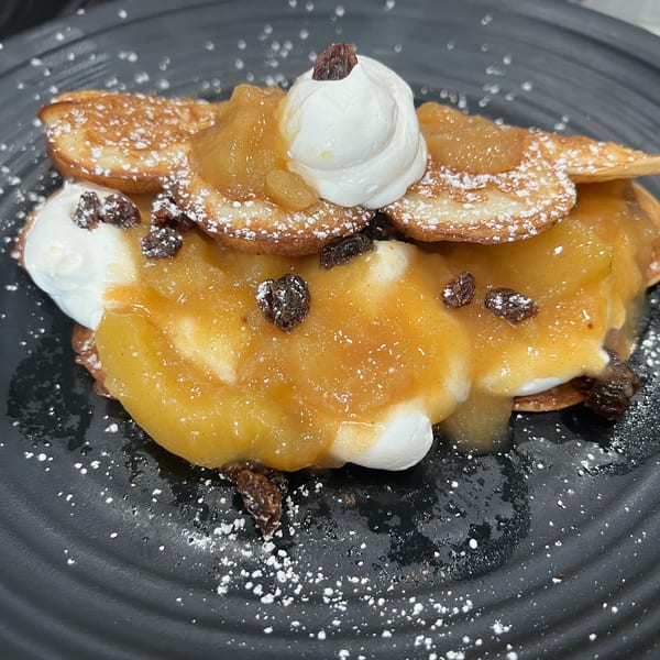 Pancakes with apple compote, whipped cream, raisins, and powdered sugar on a black plate.
