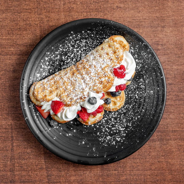 Sweet folded waffle topped with whipped cream, mixed berries, and powdered sugar on a black plate.