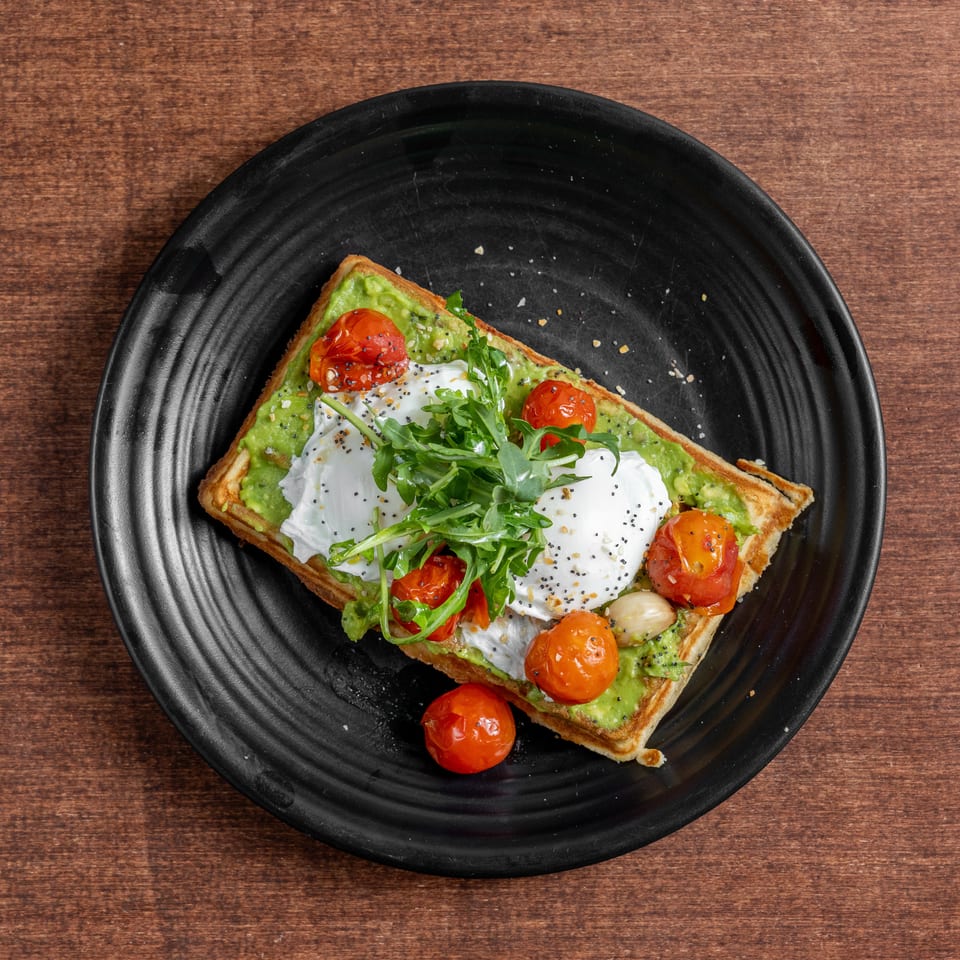 Waffle topped with avocado, poached eggs, roasted tomatoes, arugula, and poppy seeds on a black plate.