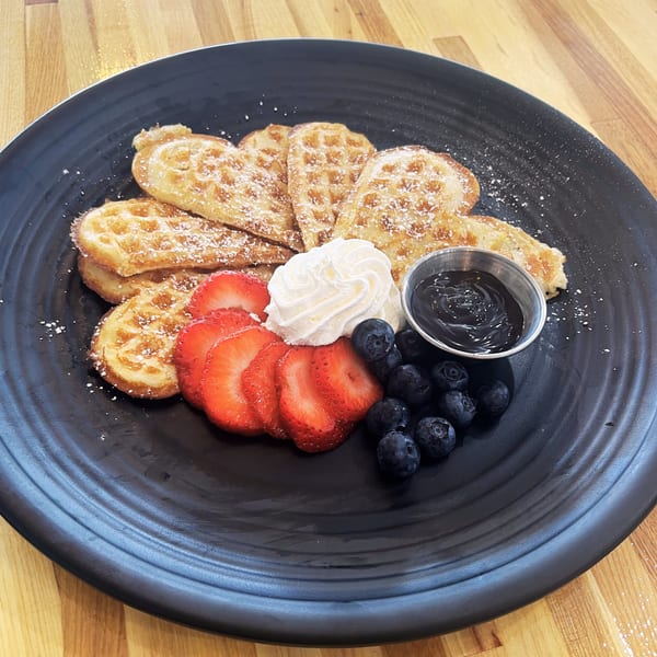 Heart-shaped waffles with powdered sugar, sliced strawberries, blueberries, whipped cream, and chocolate sauce.