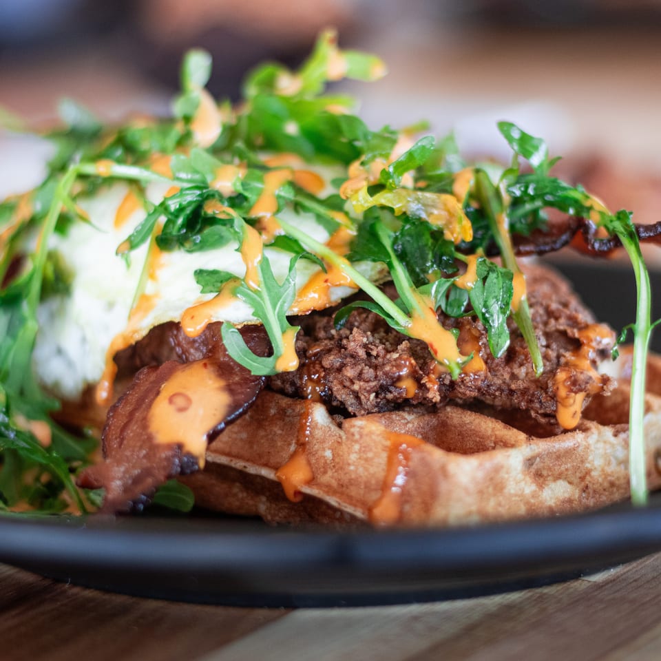 Chicken and waffles with bacon, a fried egg, arugula, and spicy sauce on a black plate.