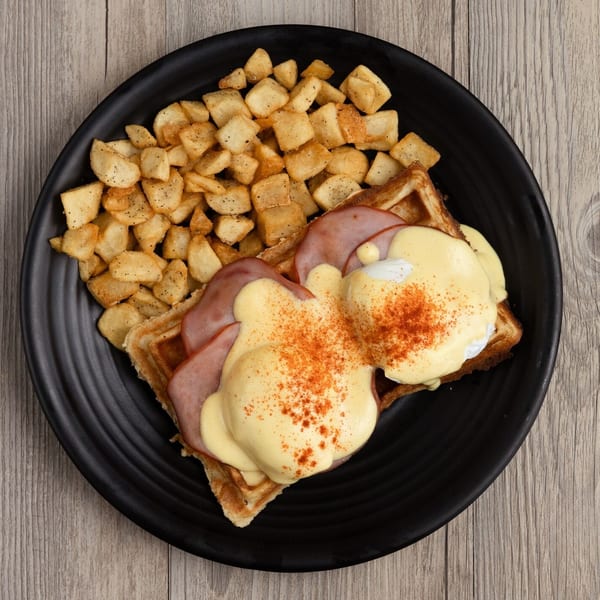 Eggs Benedict on waffles with ham and hollandaise sauce, served with home fries.
