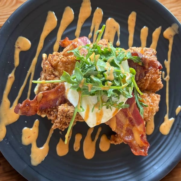 Crispy fried chicken, bacon, poached egg, and arugula drizzled with sauce on a black plate.