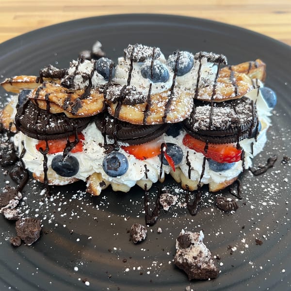 Waffle dessert topped with whipped cream, strawberries, blueberries, Oreo cookies, chocolate drizzle, and powdered sugar.