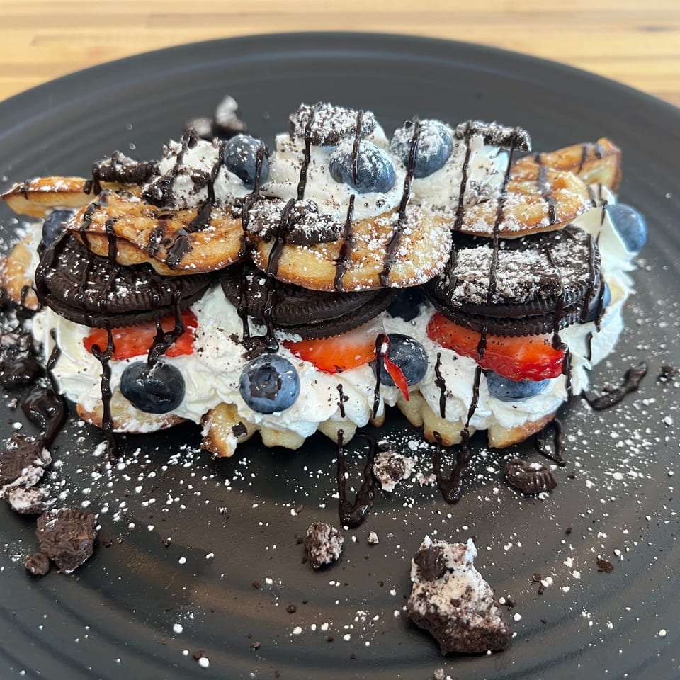 Waffle dessert topped with whipped cream, strawberries, blueberries, Oreo cookies, chocolate drizzle, and powdered sugar.